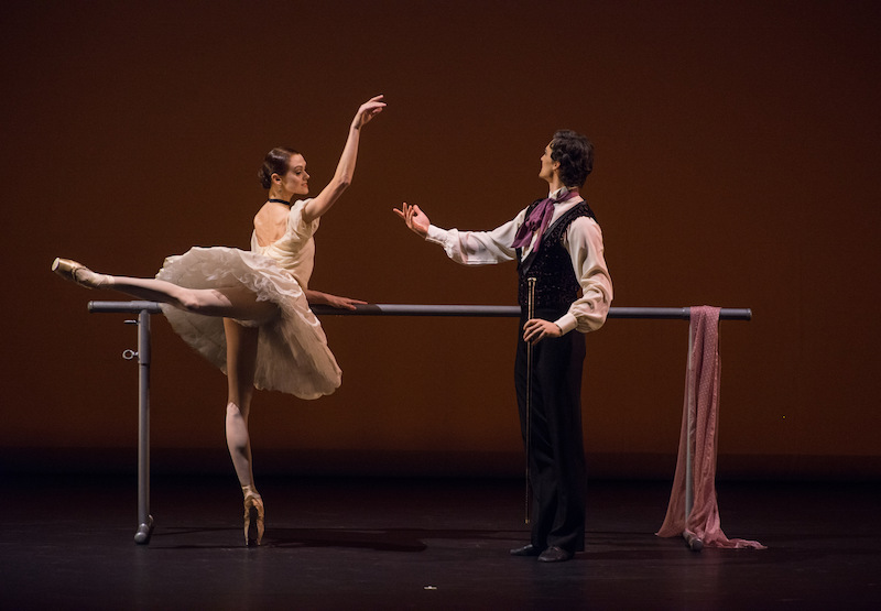 Lopatkina holds onto a ballet barre as she balances en pointe in attitude her partner gestures his hand to her.